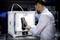A man removes a black object from a 3D printer in a laboratory. The printer is white and electronically illuminated.