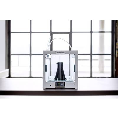 A 3D printer on a table is printing a black object. In the background, there is a large window with plenty of daylight.