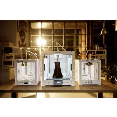 Three 3D printers on a table in a lit room. The middle printer is creating a large, black object.
