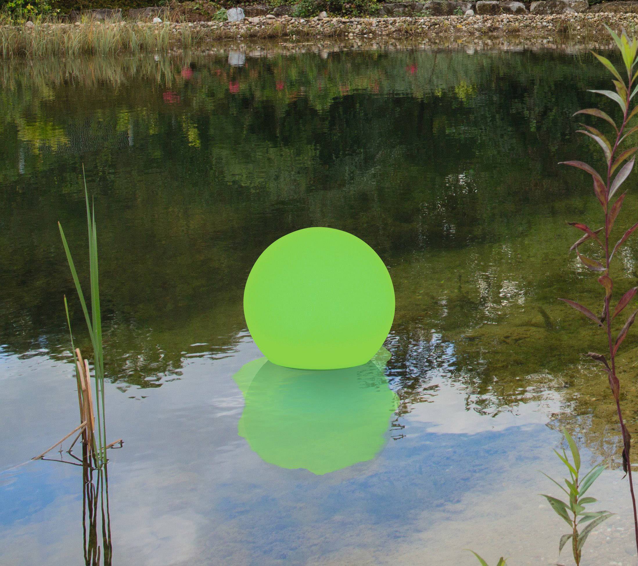 A luminous green ball floats on a clear pond, surrounded by plants and trees reflecting in the water.