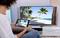 A person is looking at a beach with palm trees on a television screen, while holding a tablet displaying the same image.