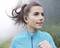A woman jogs in a blue jacket on a misty day. She is wearing wireless earphones and looks ahead with concentration.