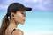 A woman on the beach wearing a black cap and wireless earphones looks out to sea. The background shows light blue water and sky.