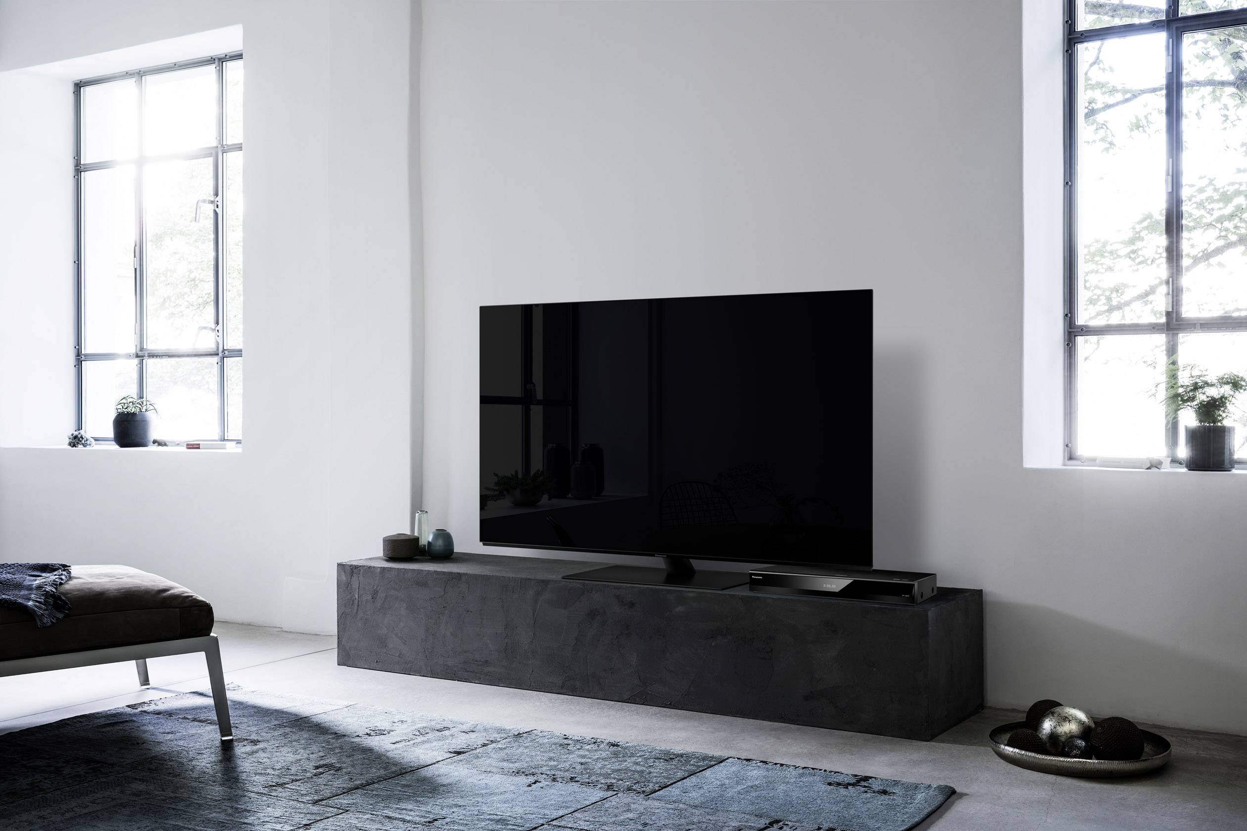 A modern living room with a large flat-screen television on a low, grey table. Large windows allow plenty of light to enter.