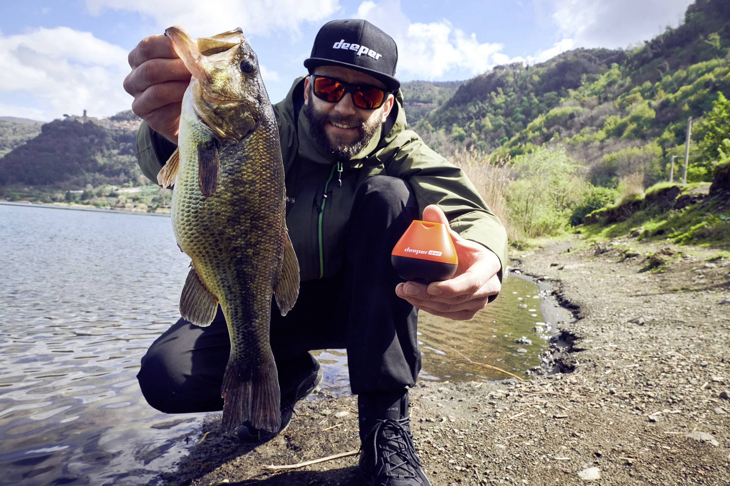 A man is holding a captured fish in his right hand and a small, orange object in his left hand. A lake is visible in the background.