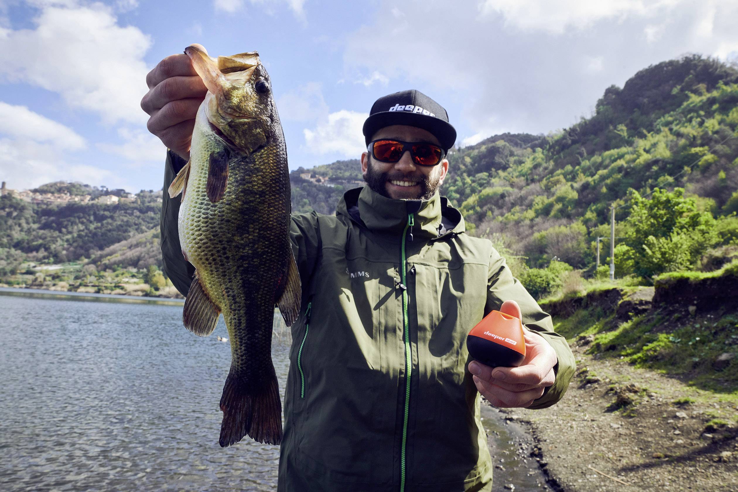 A man holds up a large fish by a lakeside and smiles at the camera. He is wearing sunglasses and a cap.