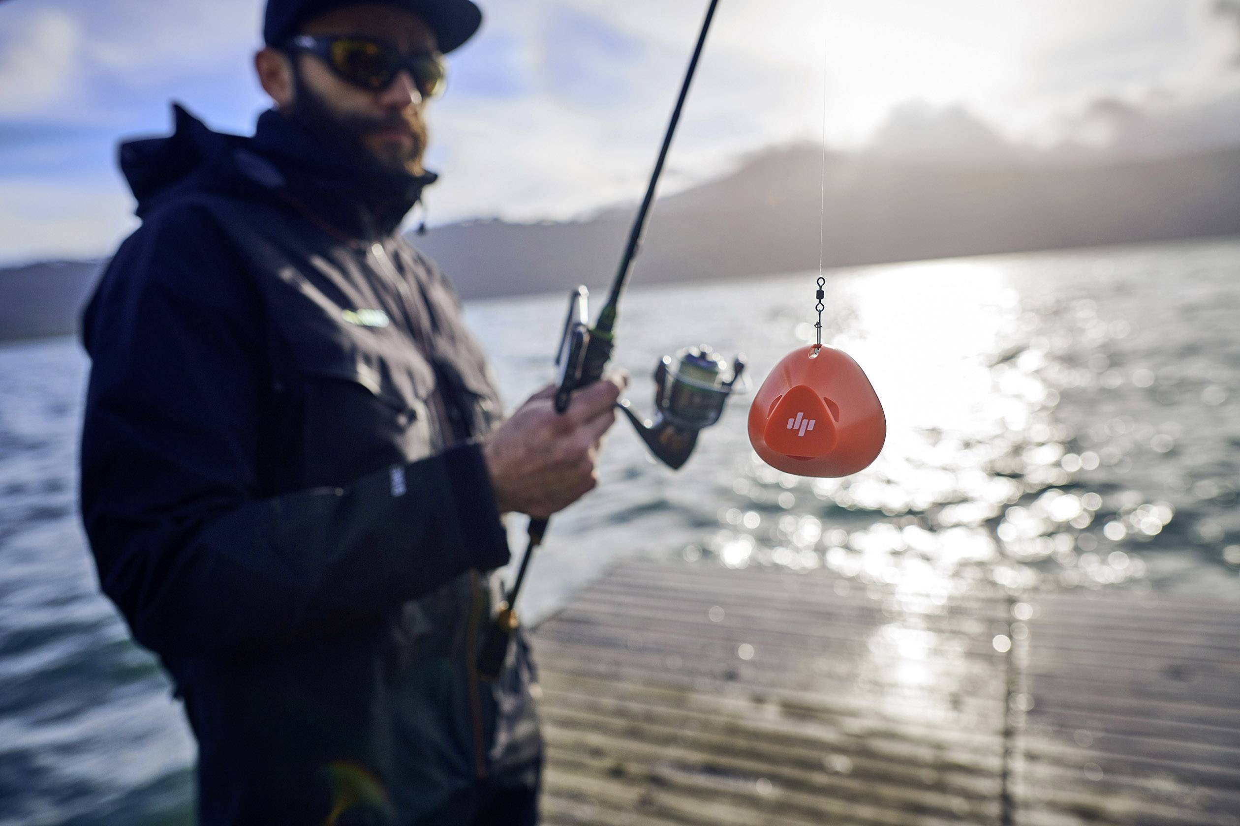 A person is fishing on a lake, holding a fishing rod with an orange device on the hook. The lake is set against a sunset backdrop.