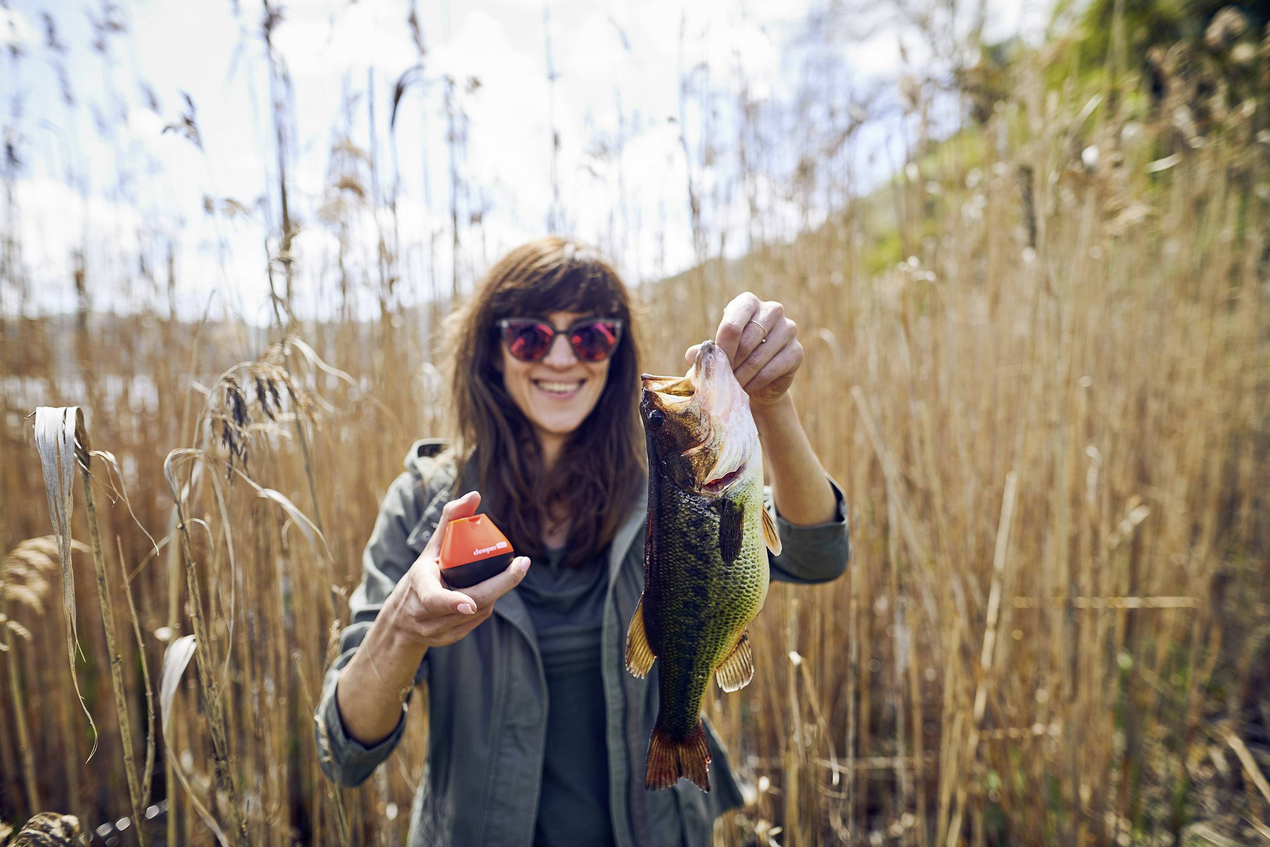 A smiling person is holding a large fish in one hand and a small device in the other, standing in a reed bed.