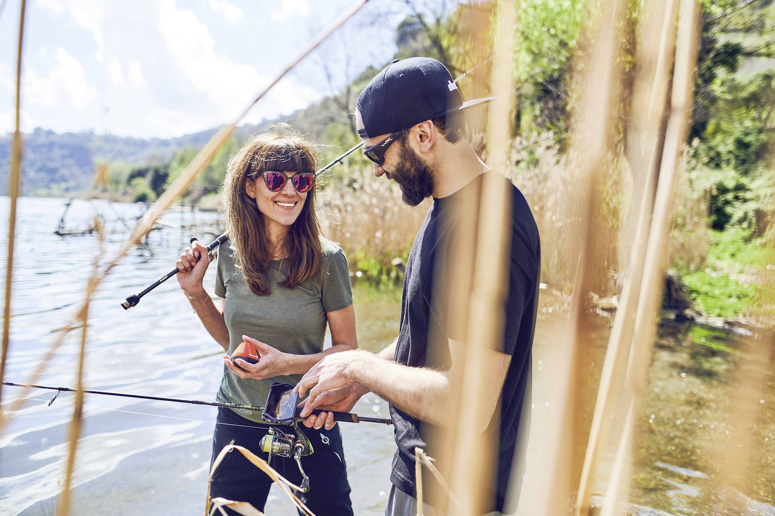 A man and a woman are fishing by a lakeside, surrounded by reeds. The woman is smiling and holding a fishing rod, while the man is pointing at something in his hand.