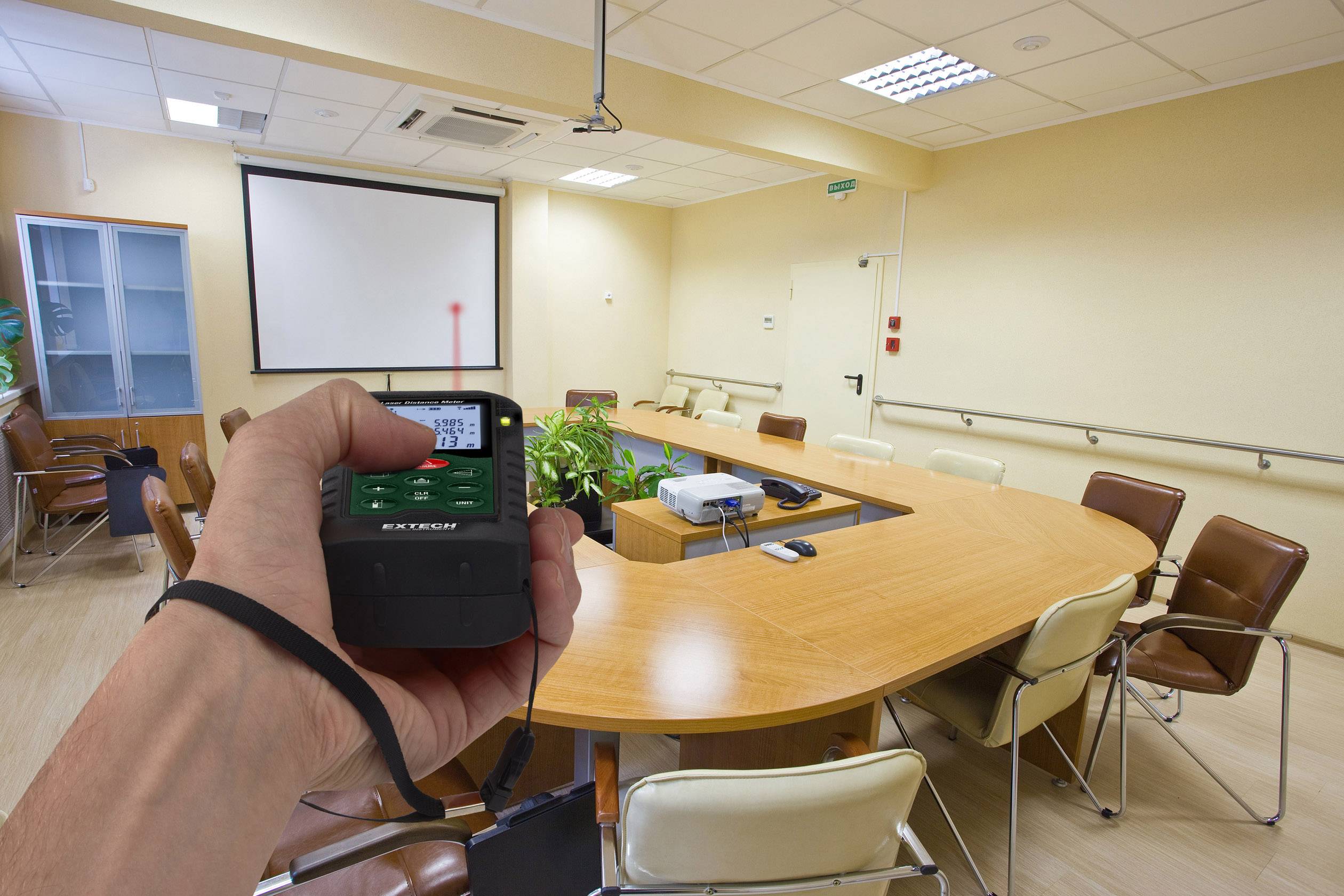 A person is holding a distance measuring device in a conference room. A red laser point is hitting the screen. An oval table with chairs stands in the room.