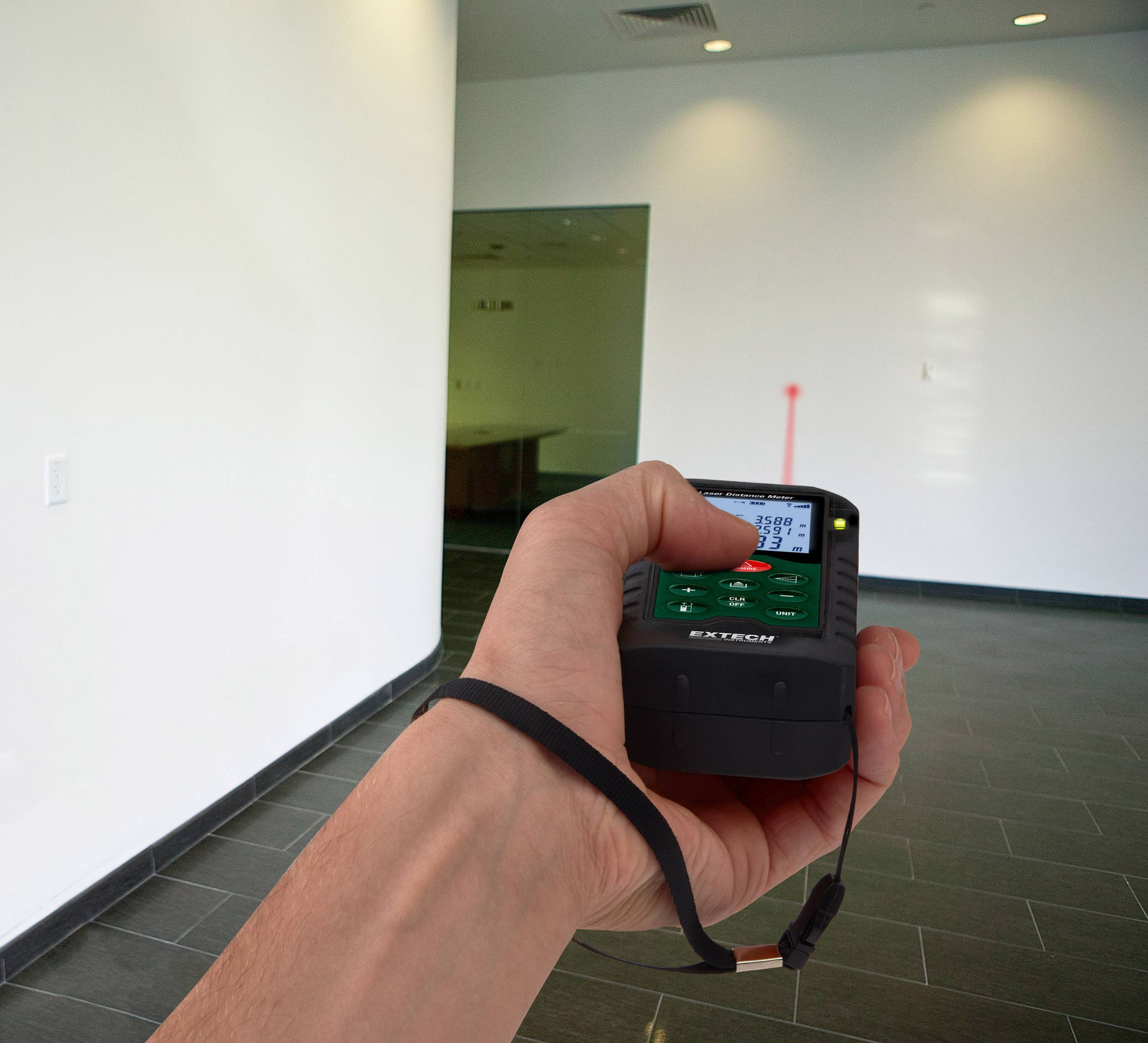 A hand holds a laser distance measurer in an empty room; a red target line indicates the measured distance.
