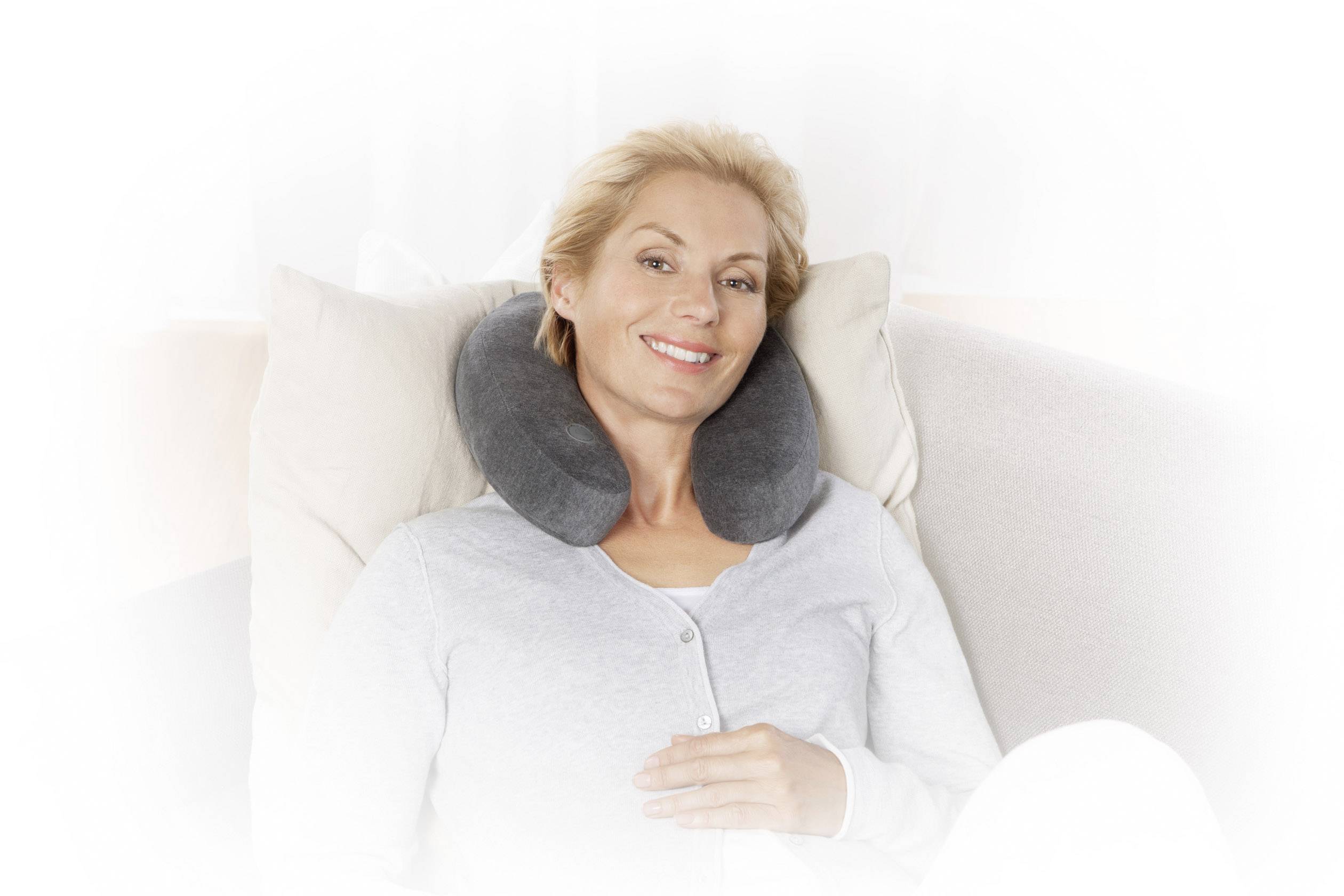 A woman is comfortably reclining on a sofa, with a grey neck pillow for support. She smiles relaxedly at the camera.