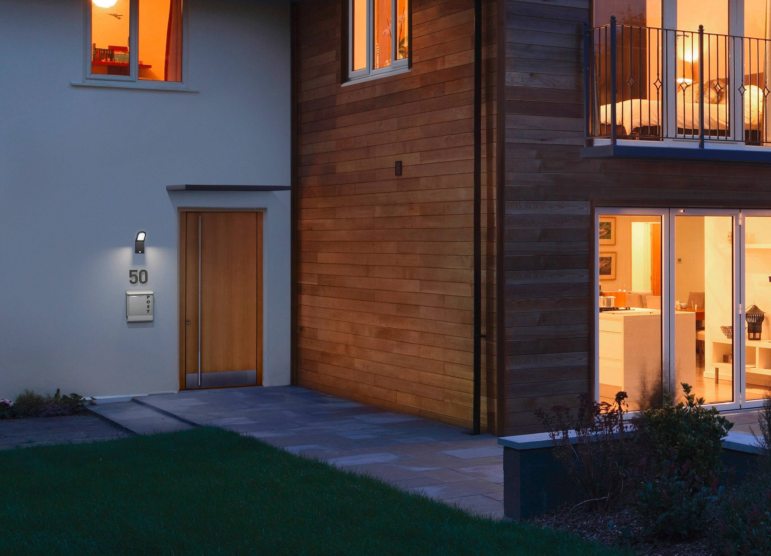 A modern building at dusk. Light glows from windows and the entrance is illuminated. House number '50' on the wall.