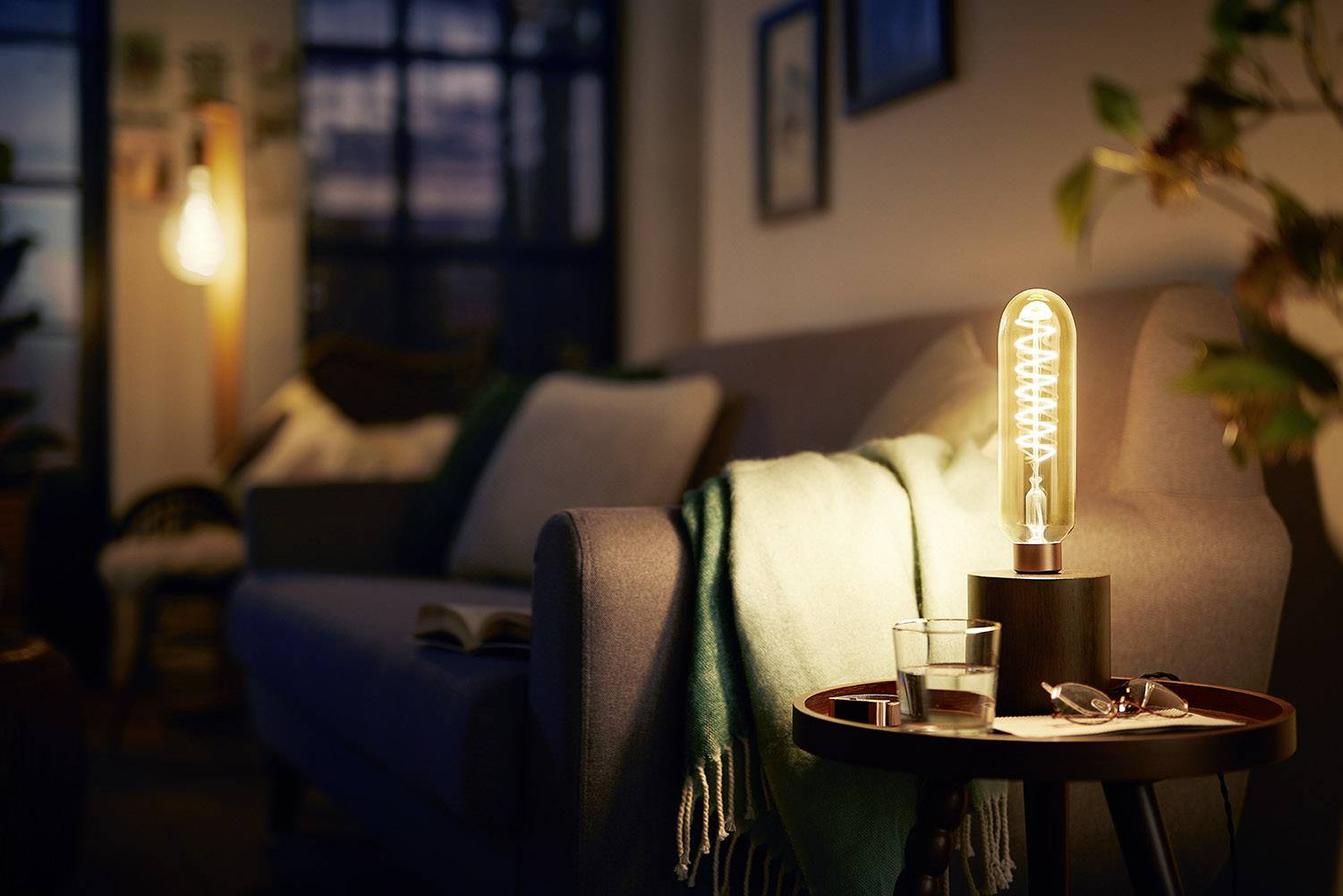 Cosy living room at dusk, with a grey sofa, throw, illuminated table lamp and glasses on a small wooden table.