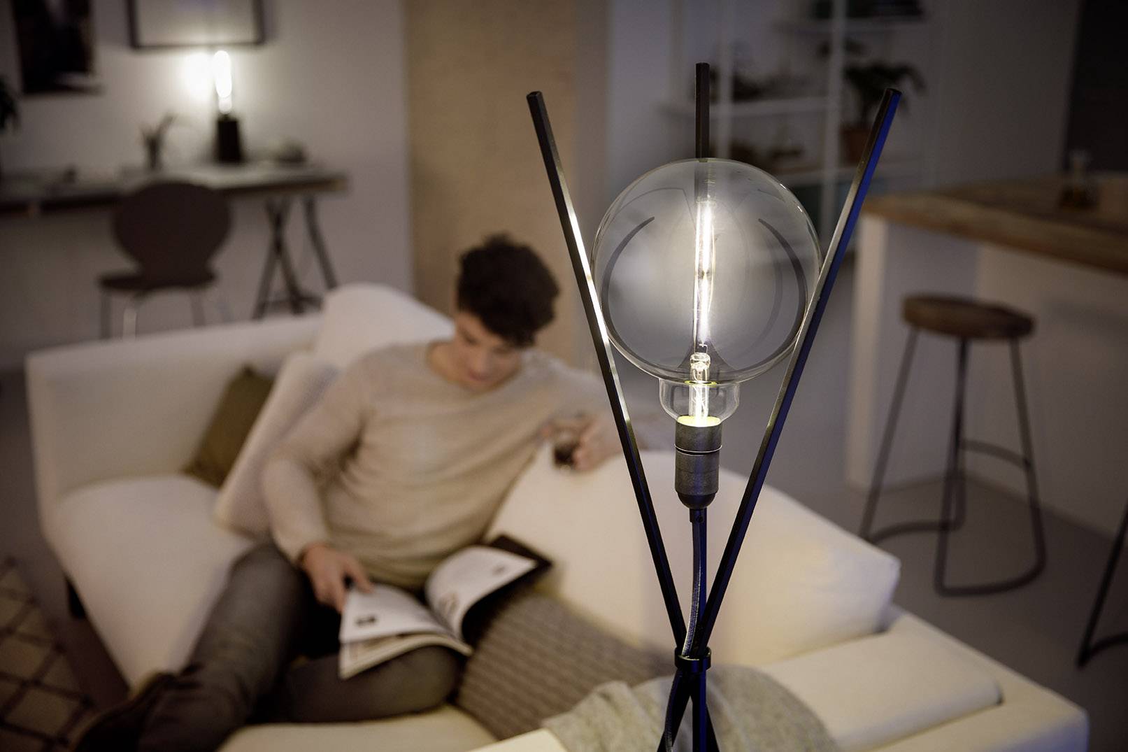 A man sits relaxed on a sofa, reading a book. In the foreground, a decorative light bulb illuminates from a stand.