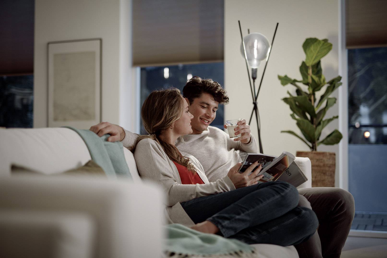 A young couple sits cosily on the sofa, reading a book together. In the background, a green houseplant stands.