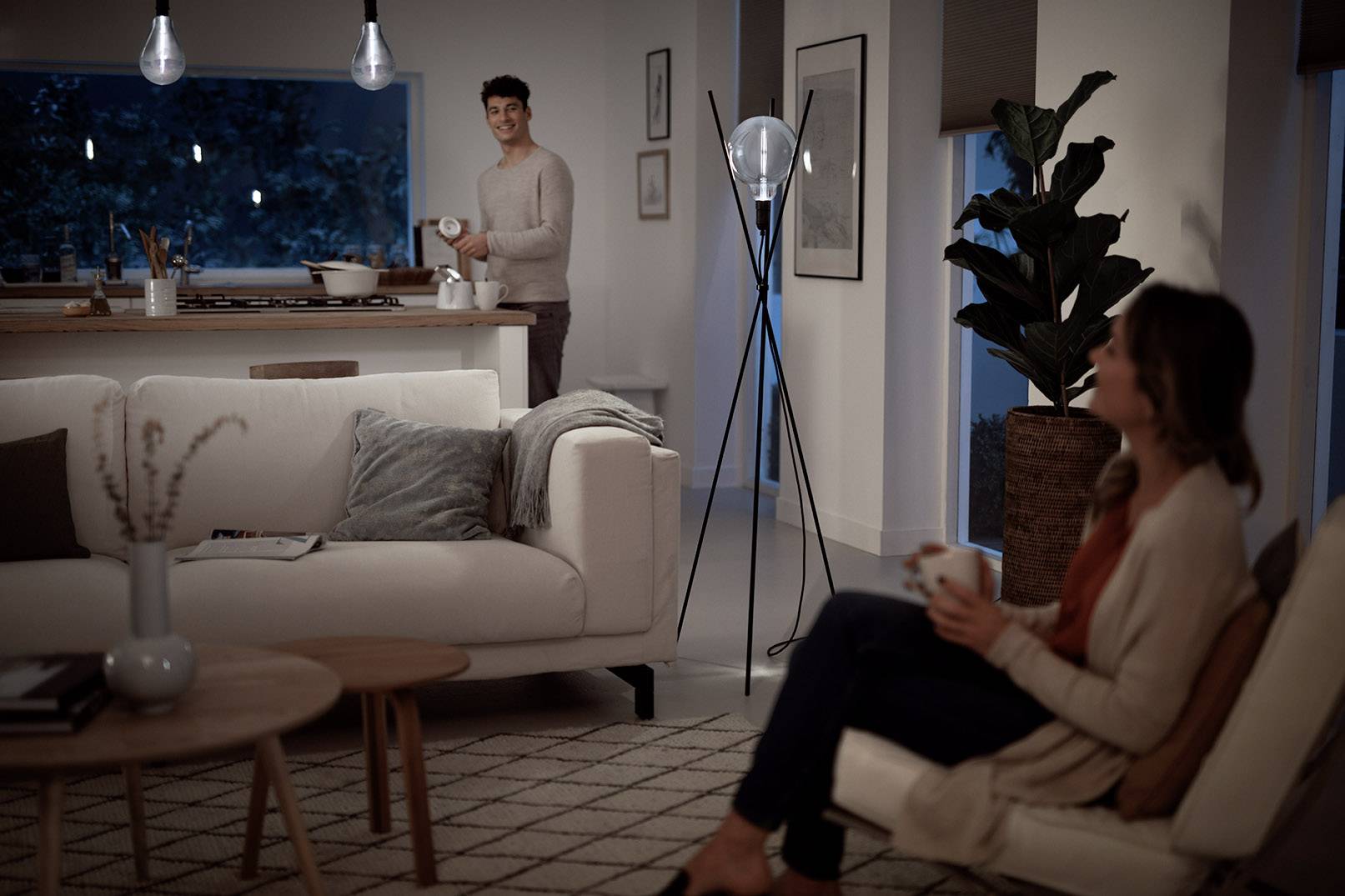 A man is standing in the kitchen with a mug, whilst a woman sits on a chair in the living room, both relaxed in a modern flat.