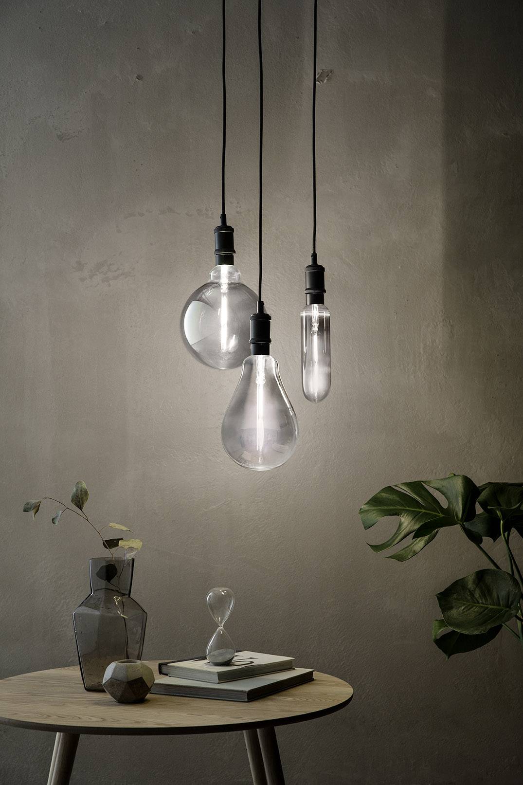 'Three modern light bulbs hang over a round wooden table with a book, vase, and plant. The scene appears minimalist and stylish.'