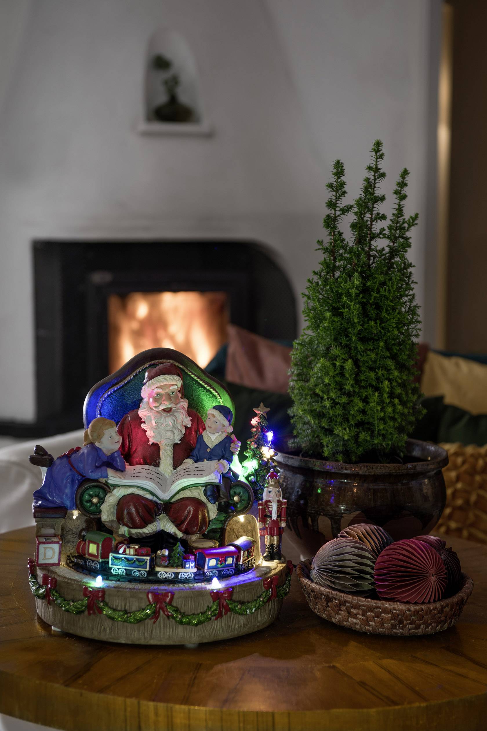 Christmas decorations on a table with an illuminated Santa Claus, miniature railway, small tree and woven bowl. A fireplace is burning in the background.