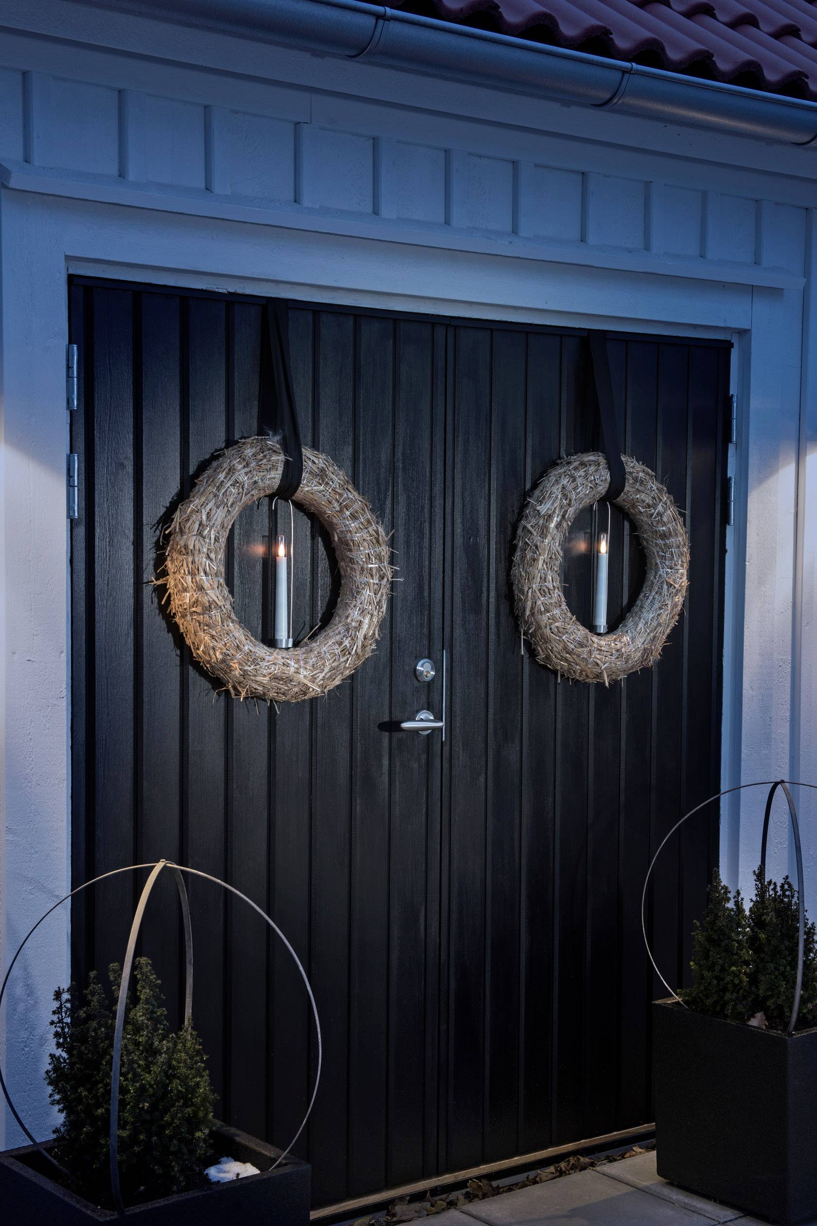 Dark wooden door with two straw-decorated wreaths, symmetrically hung. Exterior lighting casts a soft light on the door.