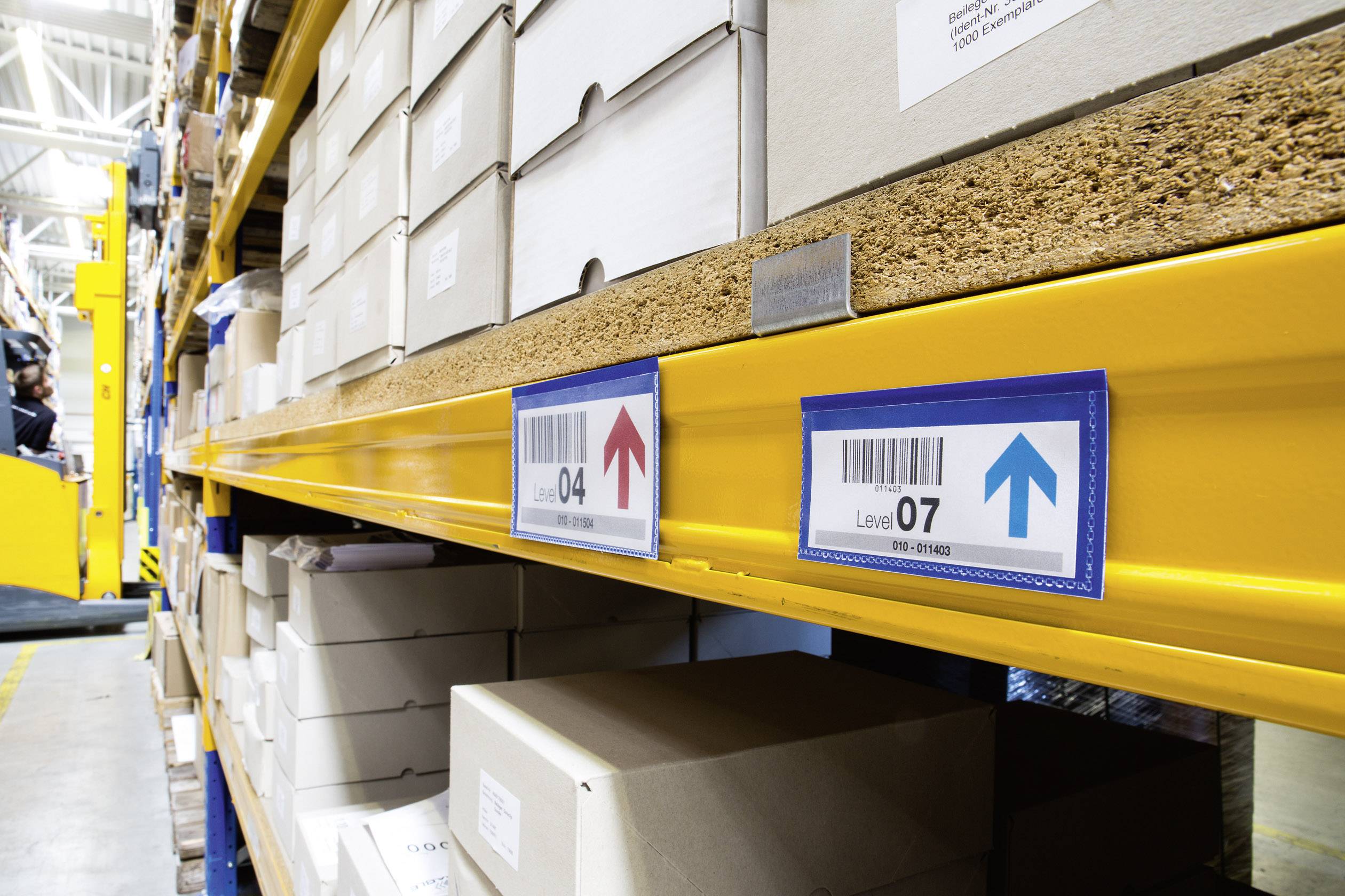 Shelving in a warehouse with cardboard boxes and arrows indicating directions on various levels. A forklift truck is in the background.
