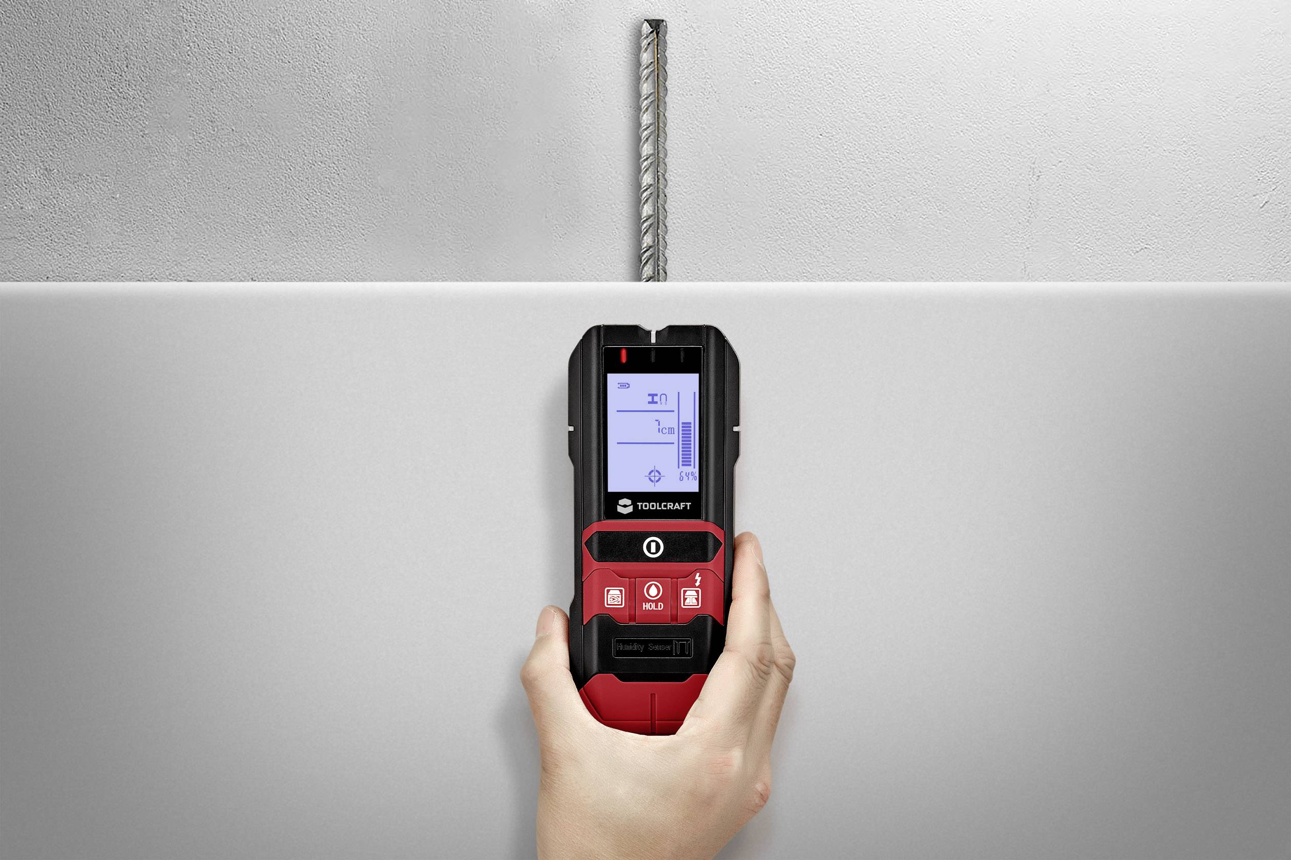 A hand holds a digital locating device showing a metal object behind a wall. The object is centrally, vertically aligned.