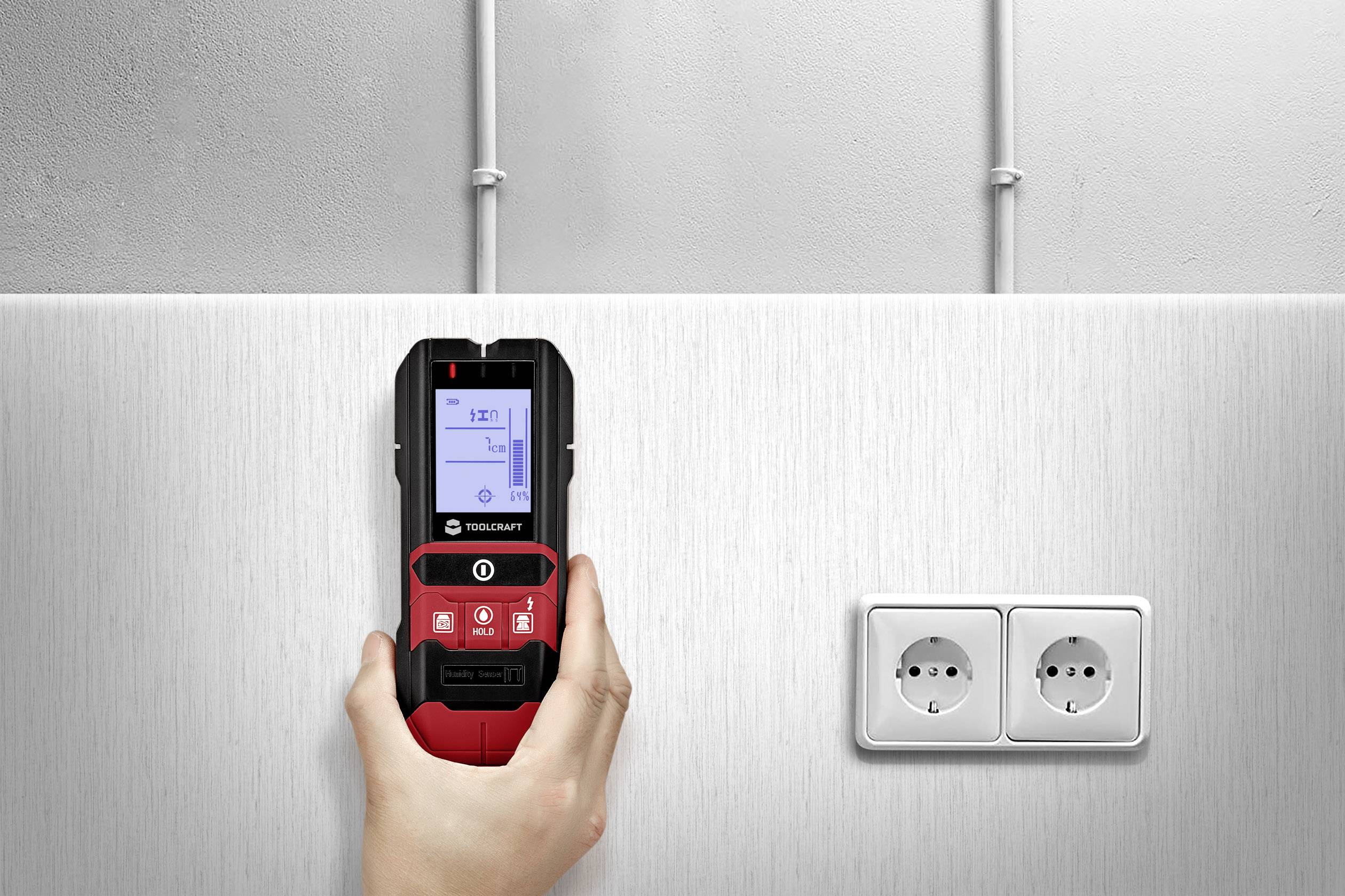 A handheld cable detection device displays information on a screen. In front of it are electrical sockets in a metal wall.