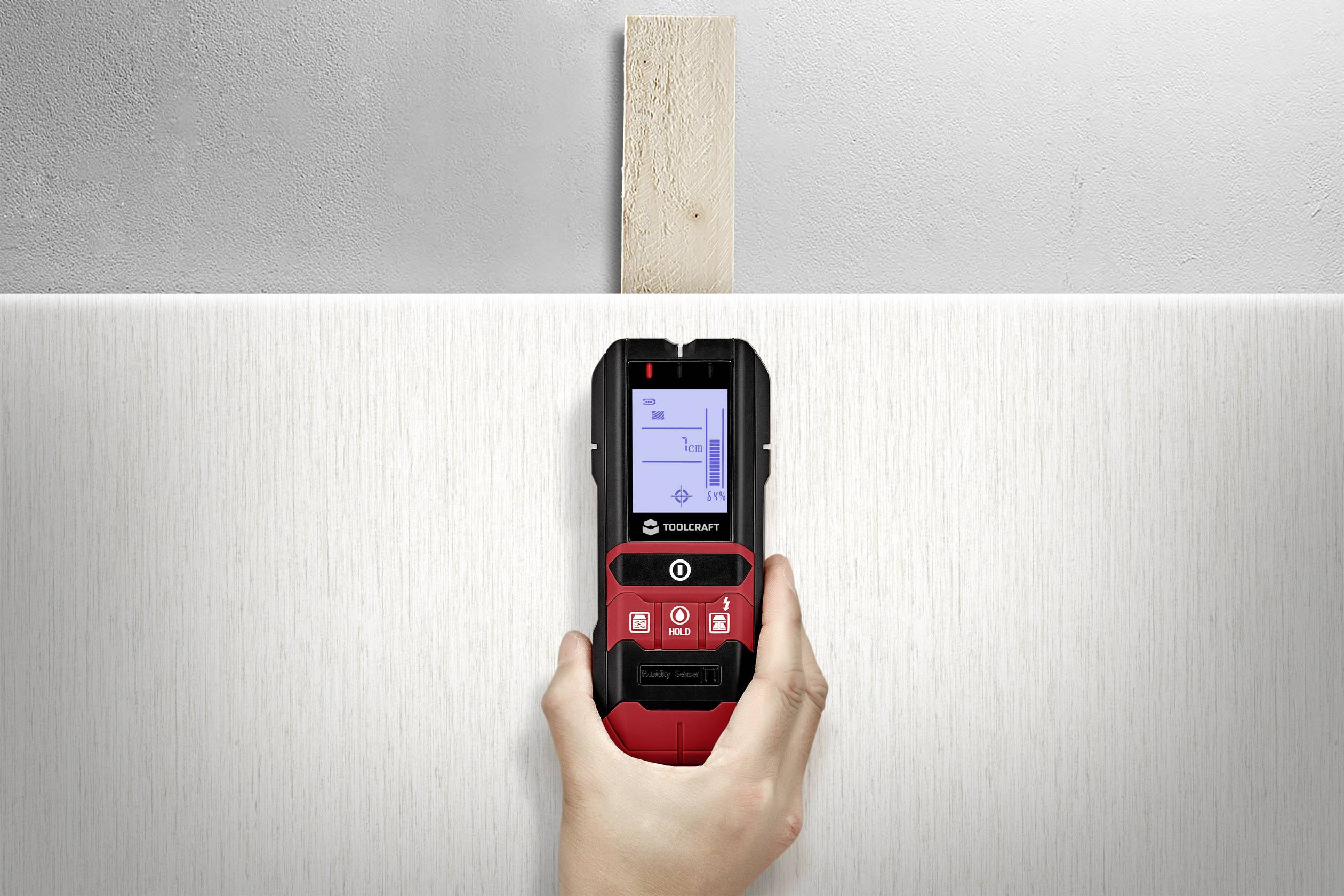 A person is holding a wall scanner device that indicates a piece of wood behind the wall. The screen displays a diagram of the detected wooden object.