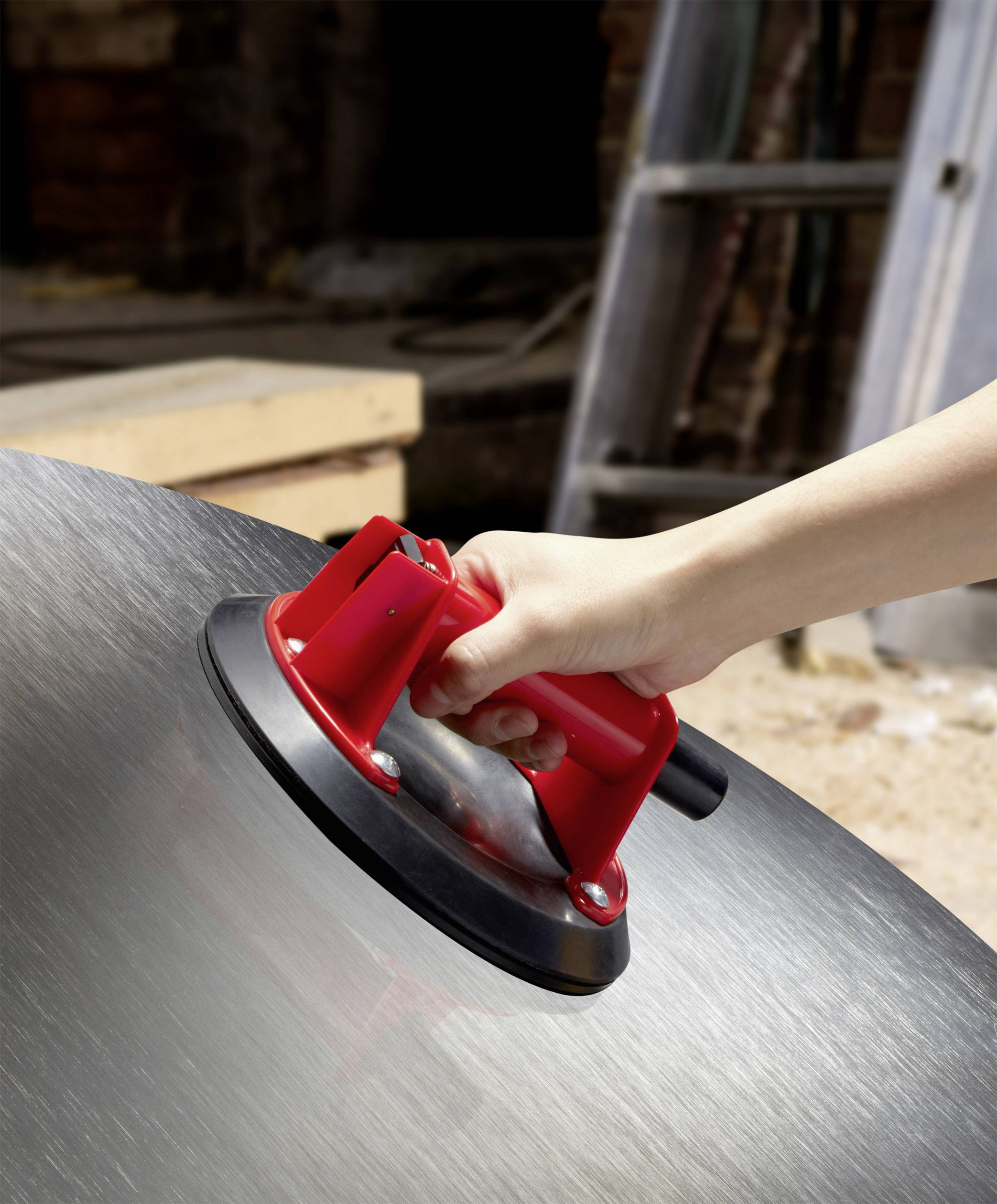 A hand is holding a red suction lifter against a large, shiny metal surface, apparently for transporting heavy or smooth objects.