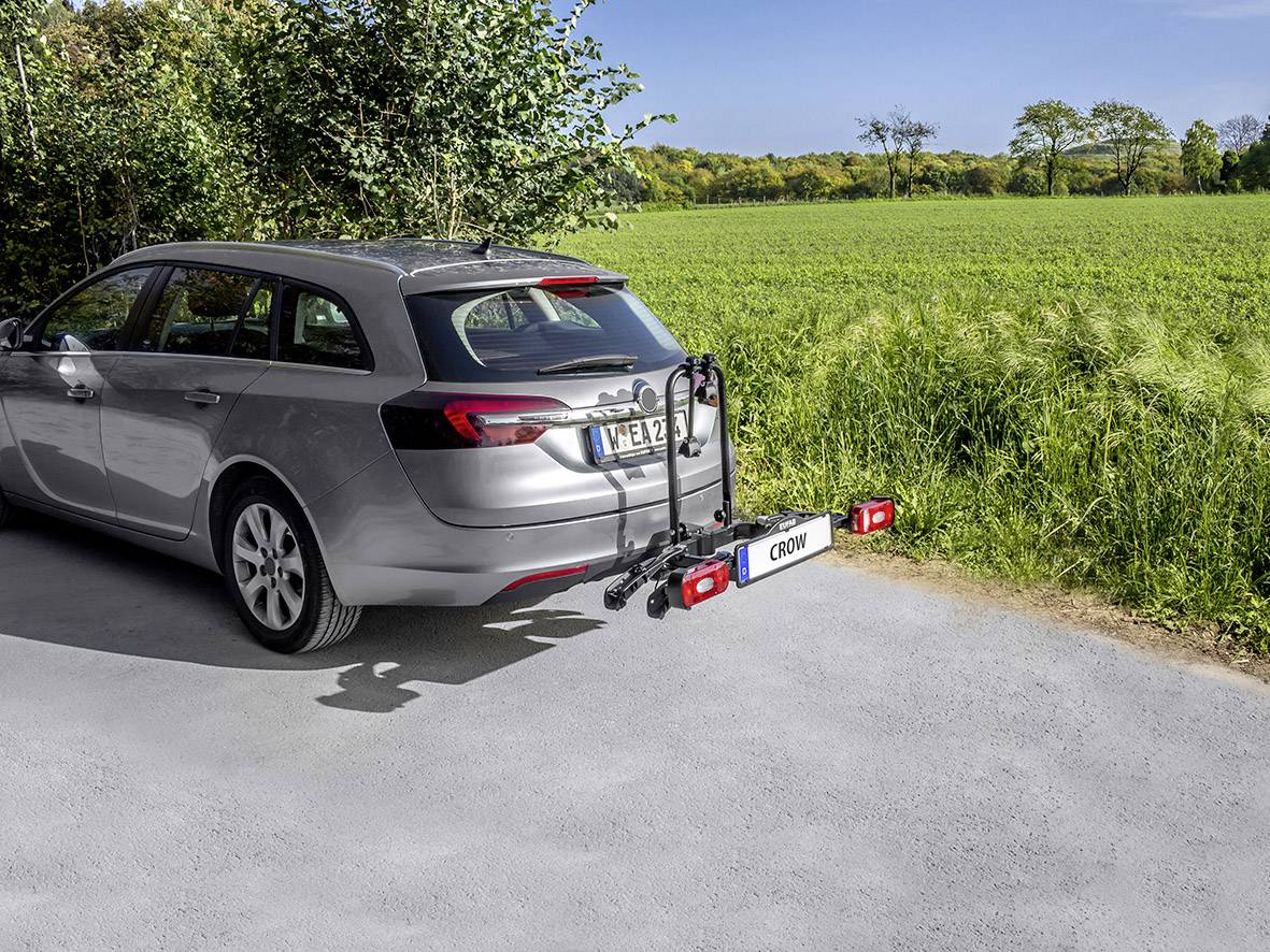 A silver estate car with a bike rack on the rear is parked on a road next to a green field and trees in the background.