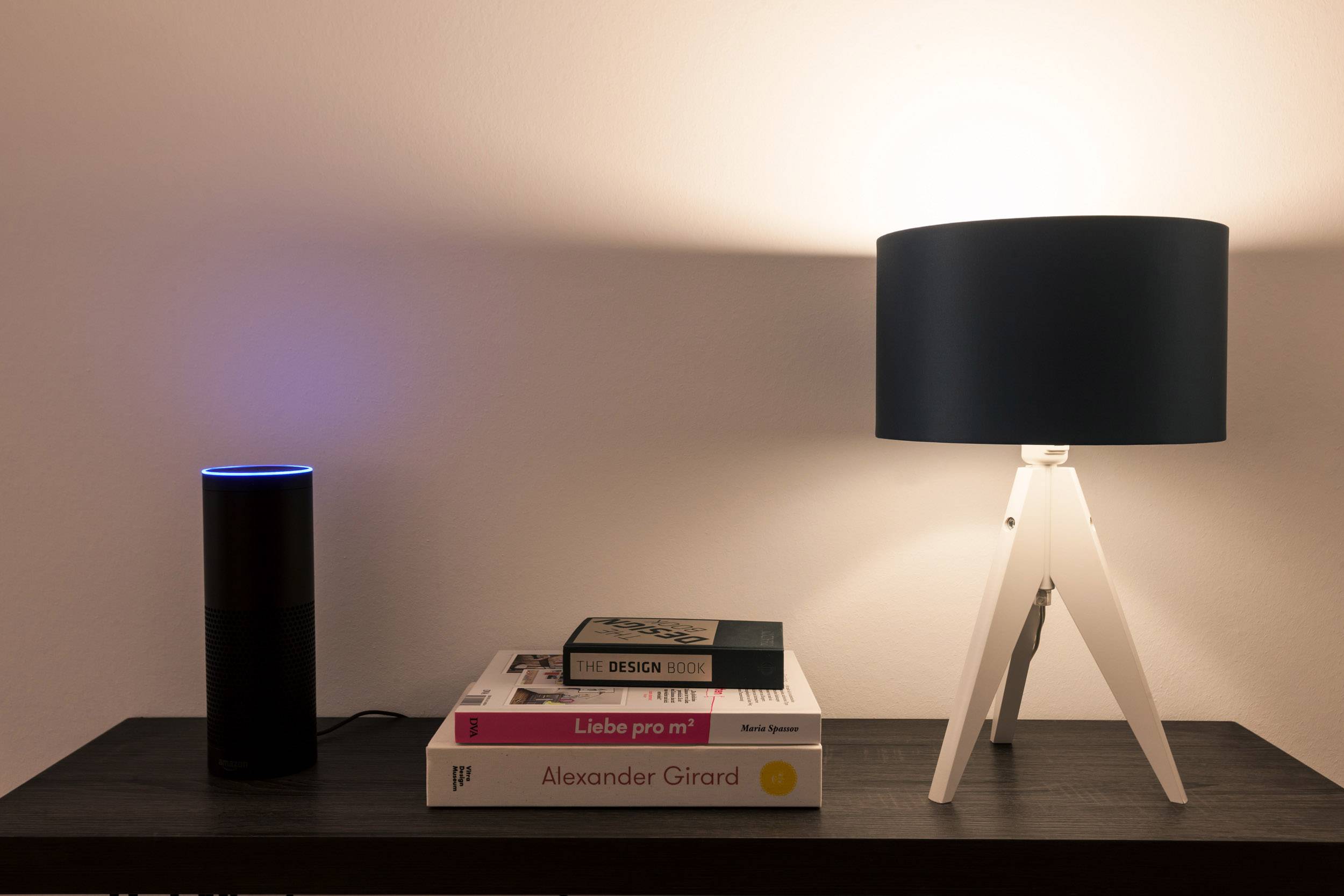 A table with a lamp on the right, which is illuminated, and a smart speaker on the left. Books are stacked in the centre.