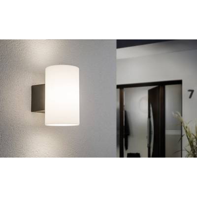A modern wall light with a cylindrical, white shade illuminates a corridor. In the background, a slightly ajar door is visible.