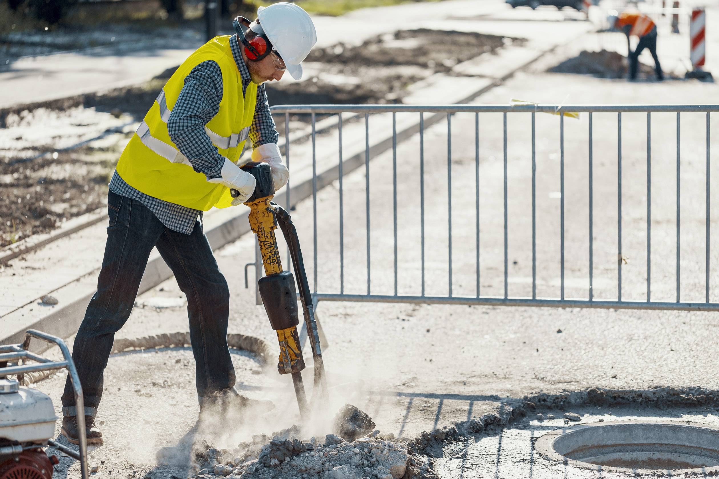 A worker in protective clothing is drilling with a pneumatic hammer on a construction site. Barriers are visible in the background.