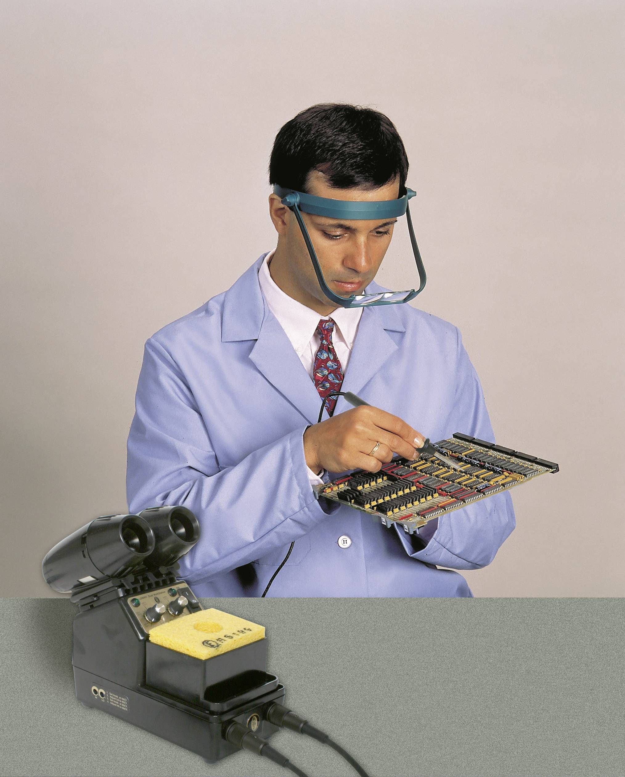 A man in a blue lab coat is inspecting an electronic circuit board. He is wearing a magnifying loupe for a more detailed examination.