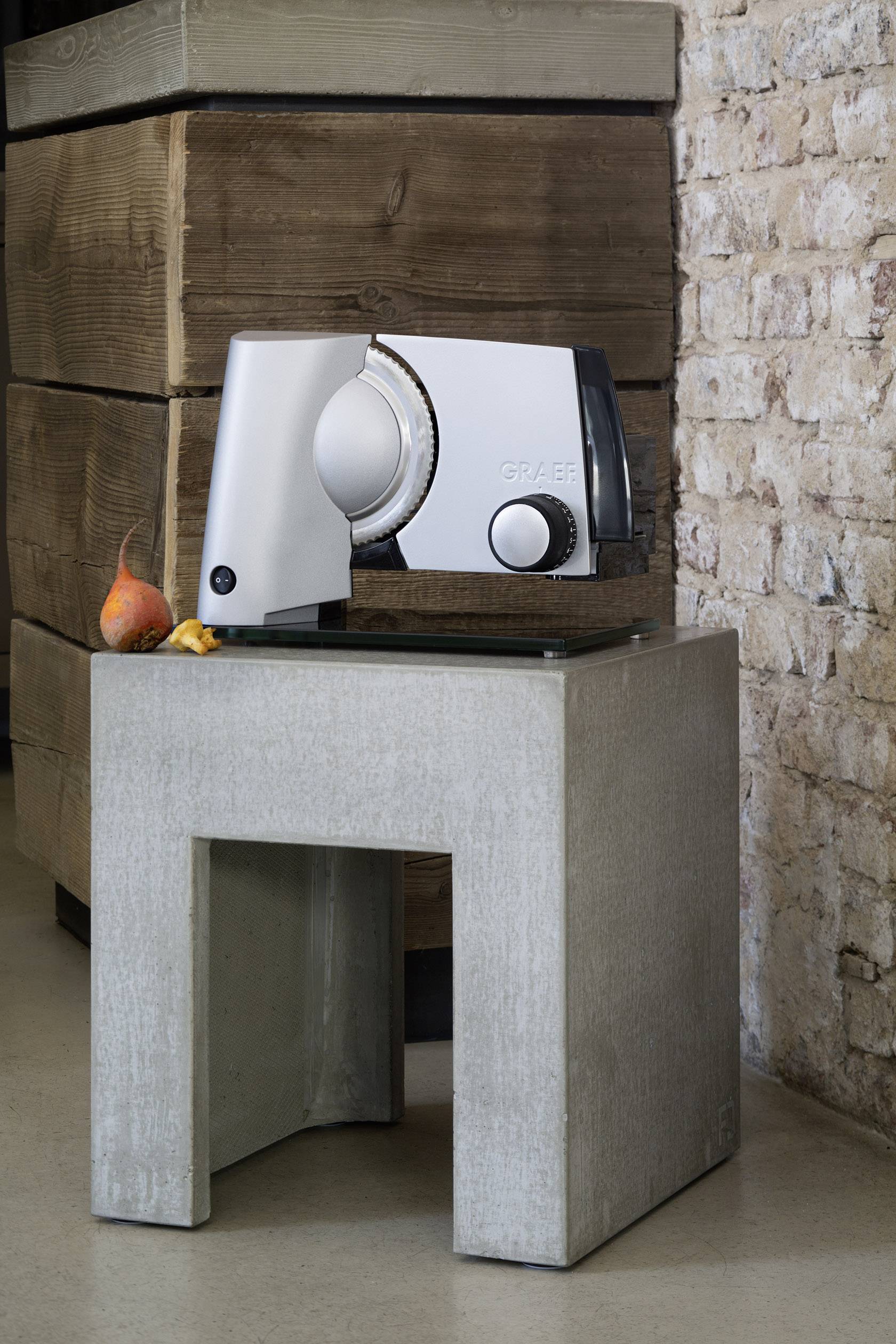 Electric all-purpose slicer on a concrete stool, next to a wall with stacked wood. An apple and a pear are lying beside it.