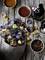 A paper bag of colourful potato crisps sits on a wooden table, surrounded by dips in bowls and drinks bottles.