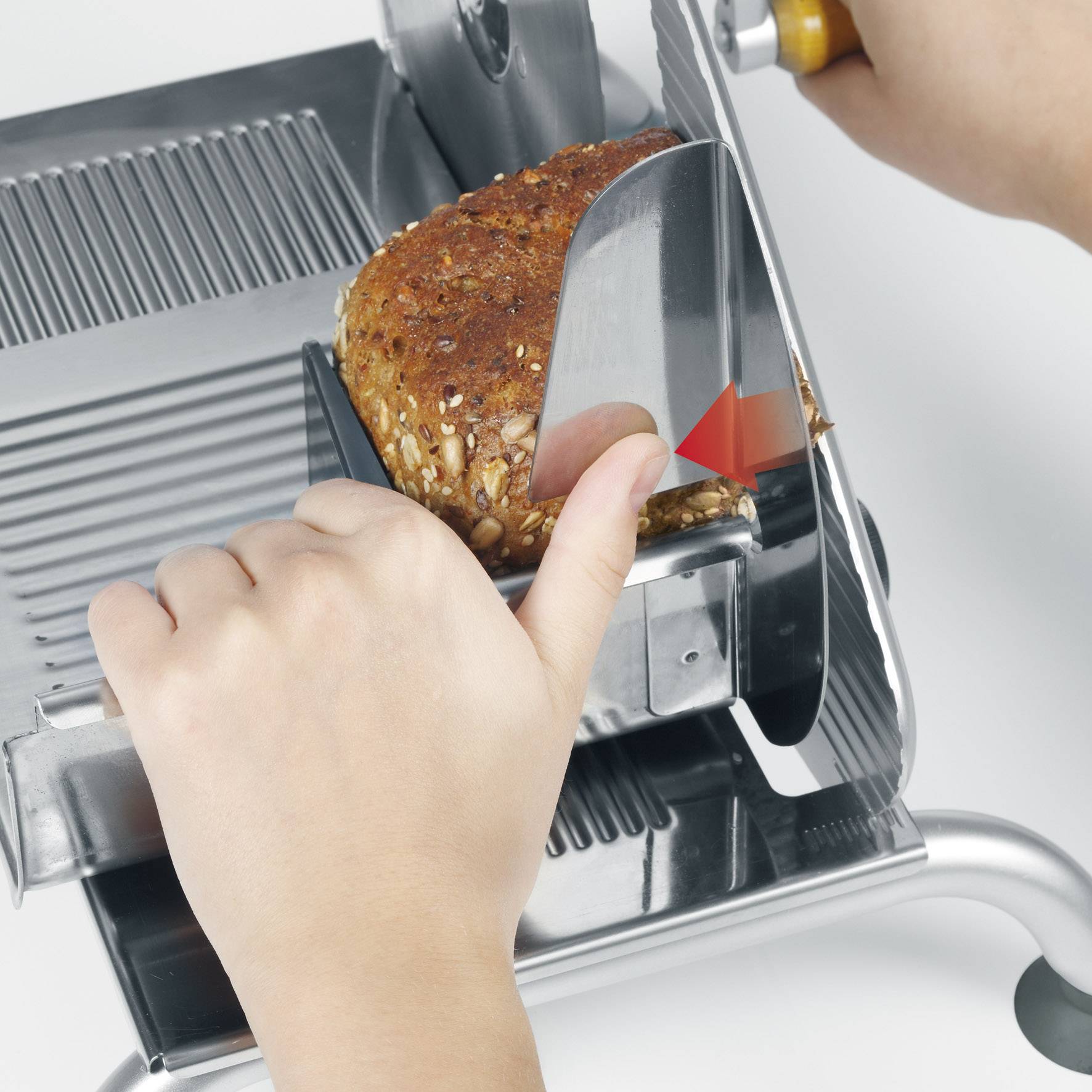 A person is cutting bread with a bread slicer. The bread has a crust with seeds. The hand is holding the bread for even slices.