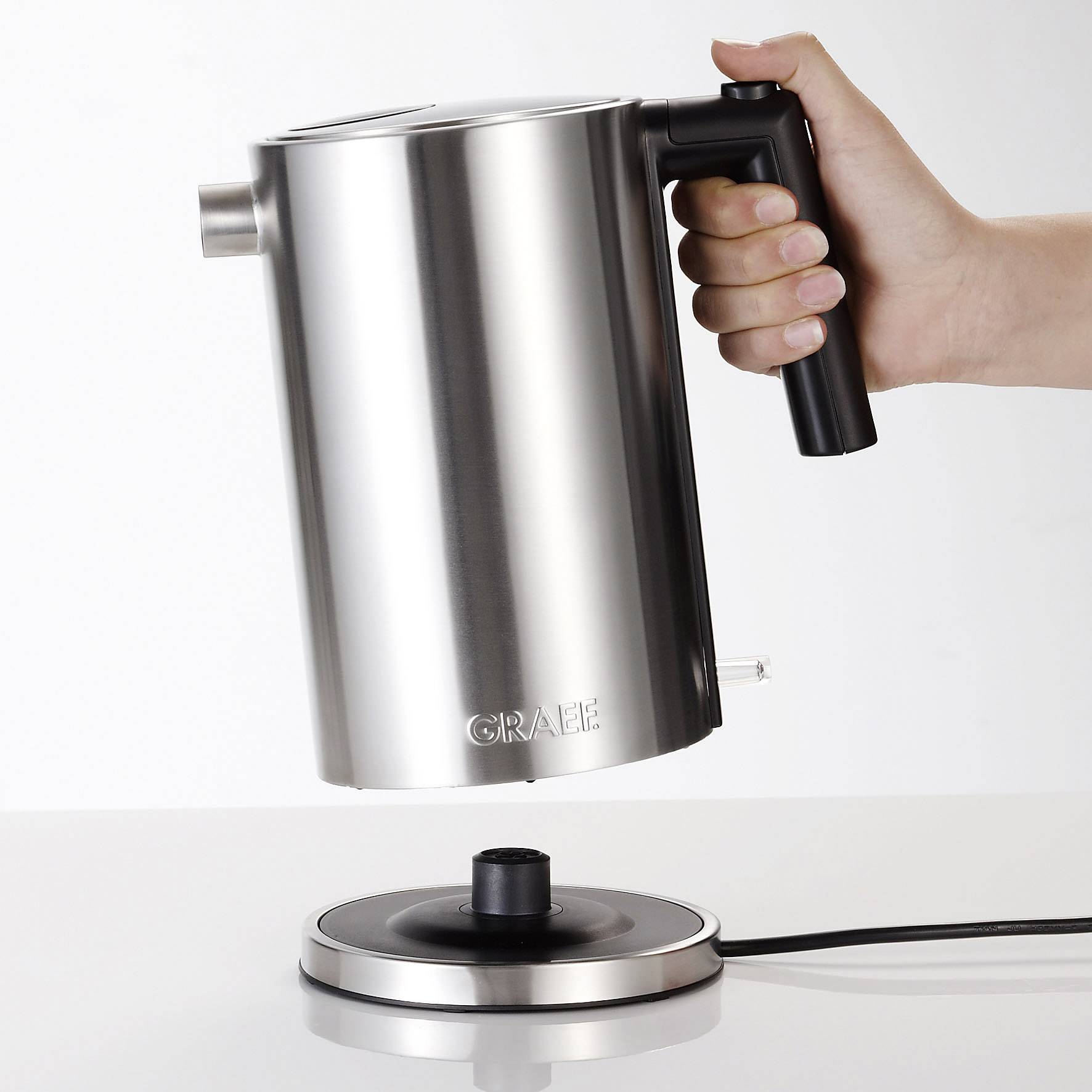 A silver kettle is being held by a hand and positioned over a round, black base.