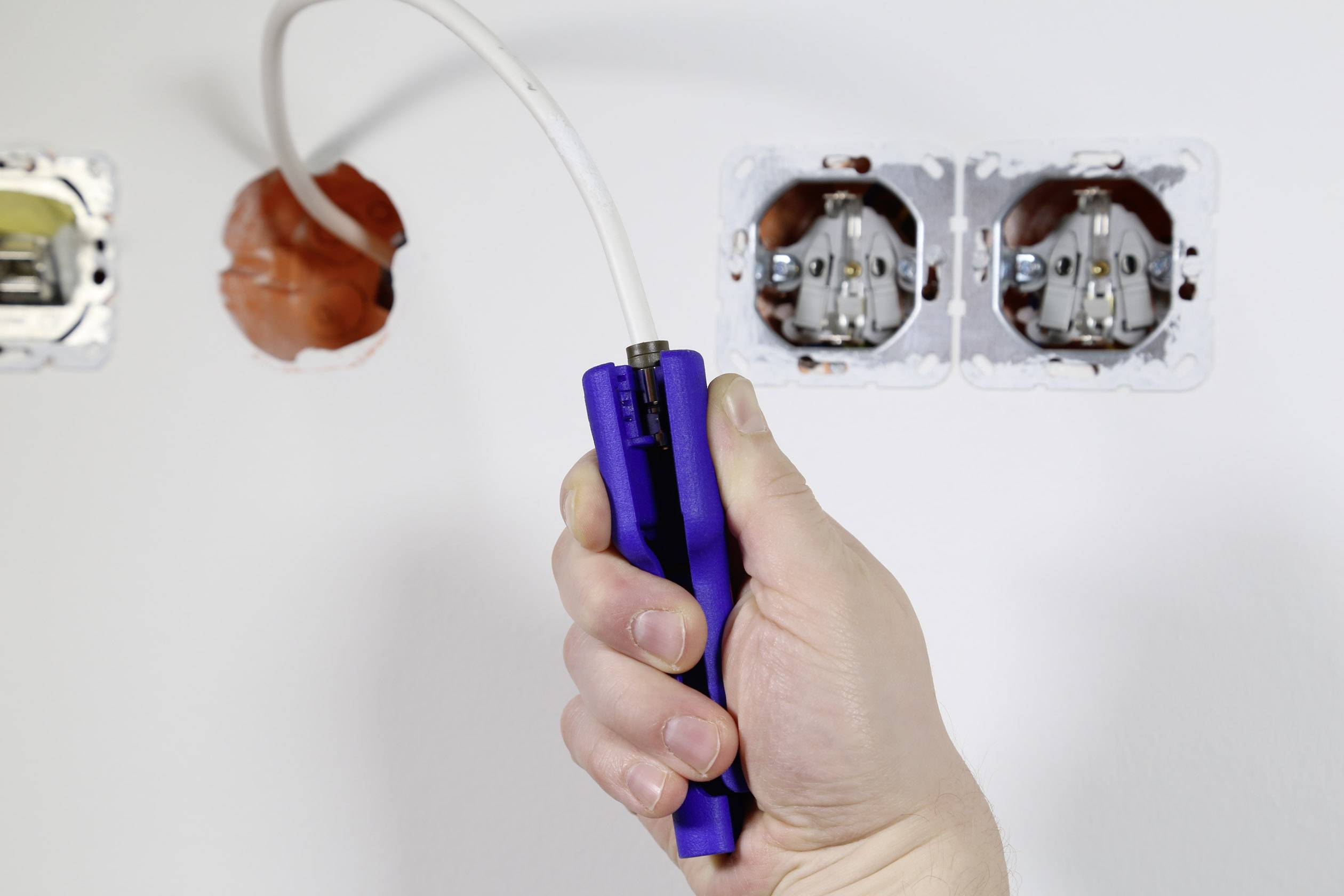 A hand is holding a blue tool that is pulling a white cable through a wall aperture. Two power sockets are visible on the right side of the image.