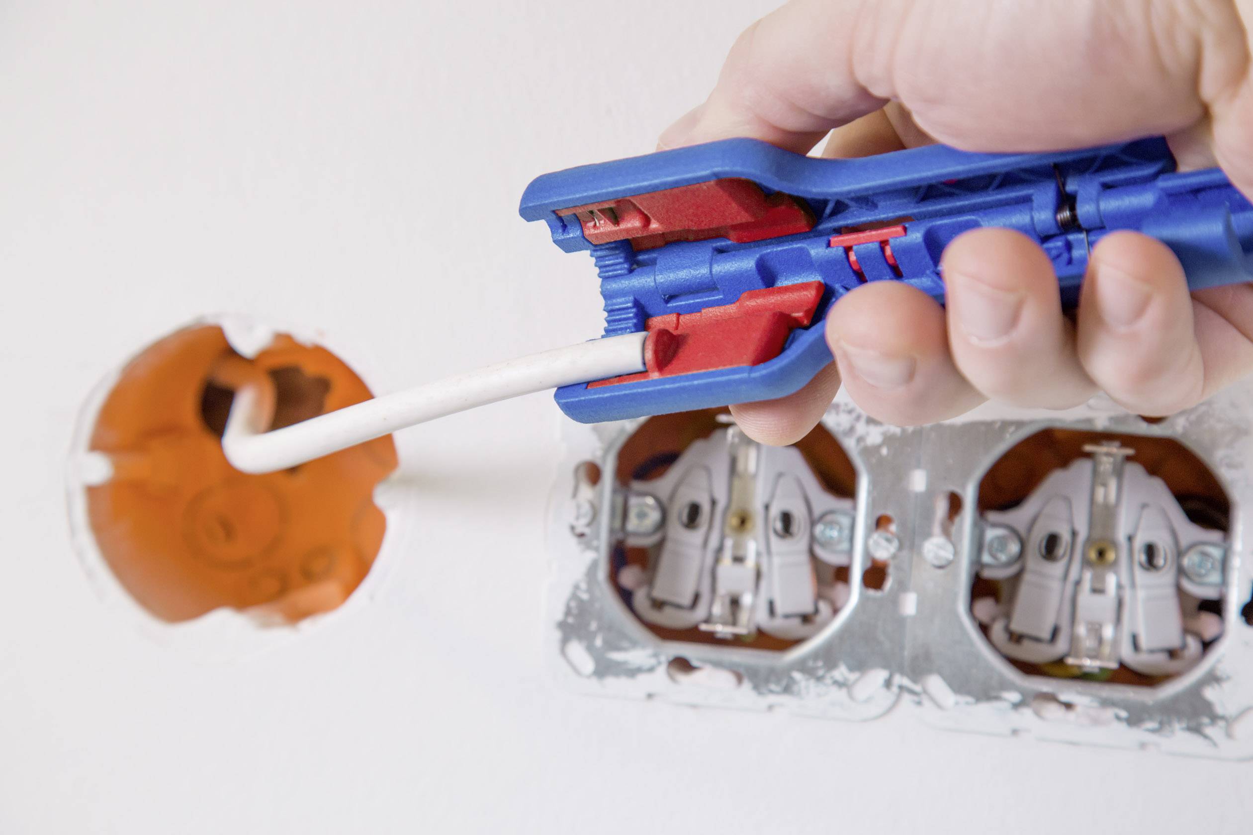 A person is using a wire stripper to strip a white cable in front of an electrical socket.