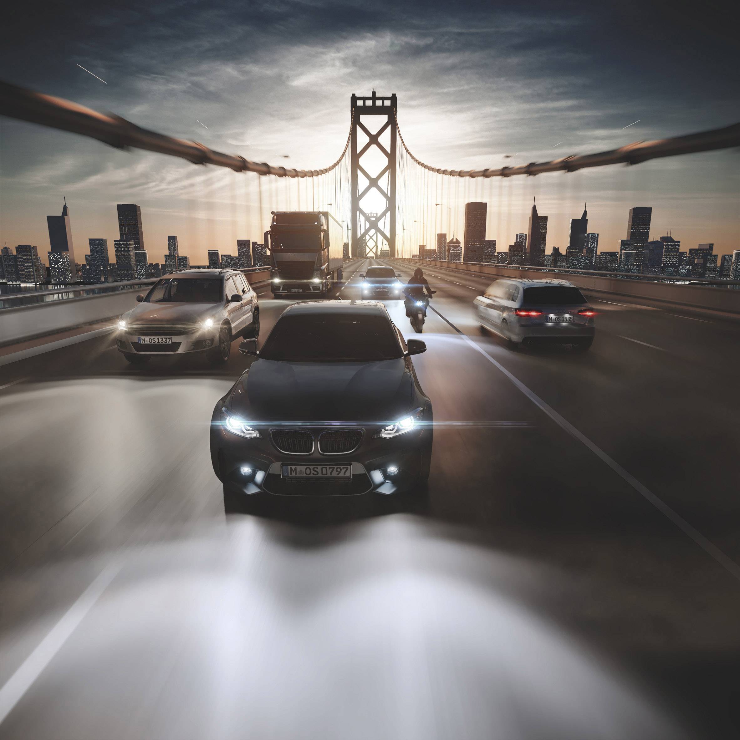 Cars drive over an illuminated bridge in a city at sunset. Skyline in the background, headlight beams visible.