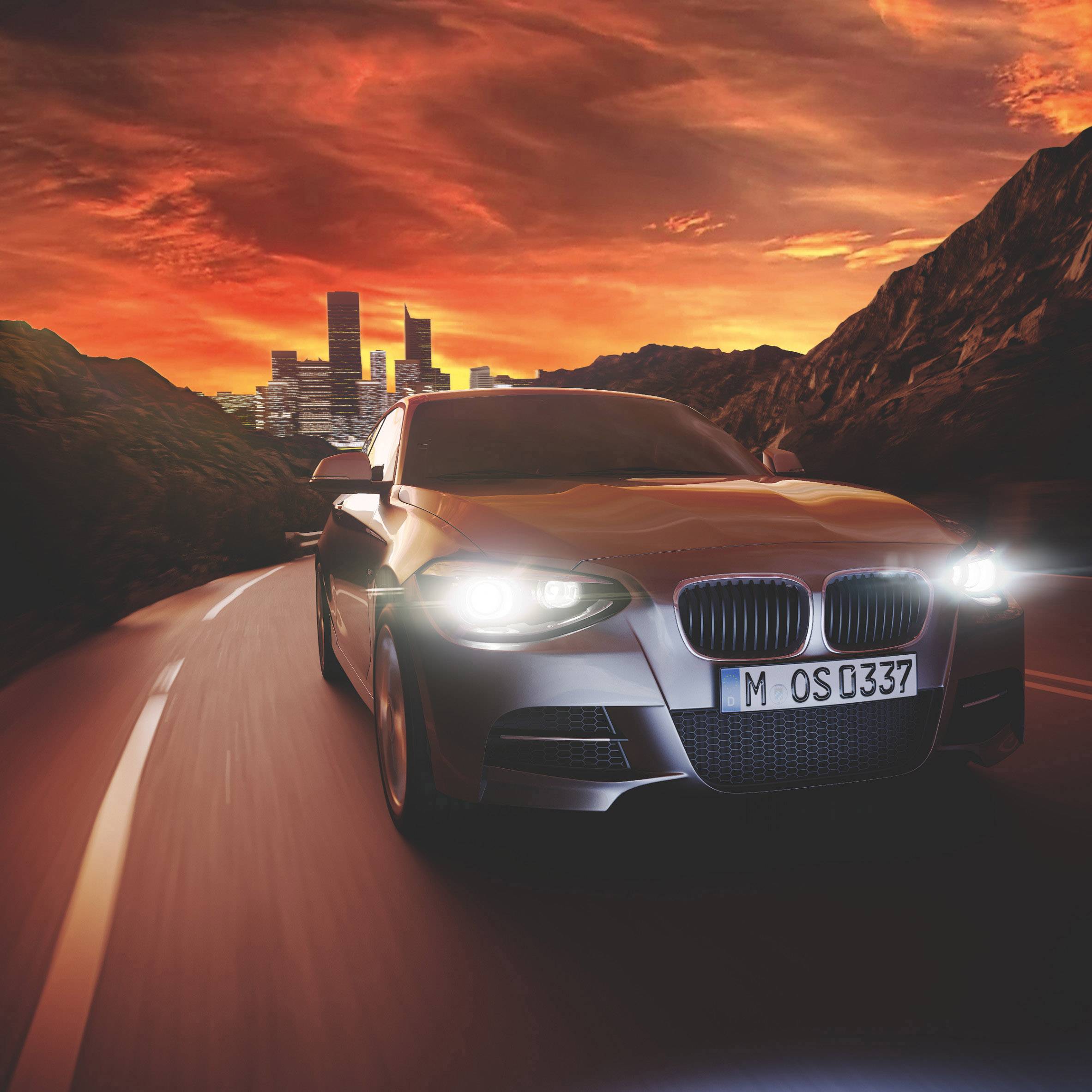 A car is driving on a road towards a city at sunset. The sky is orange, and the headlights are shining.
