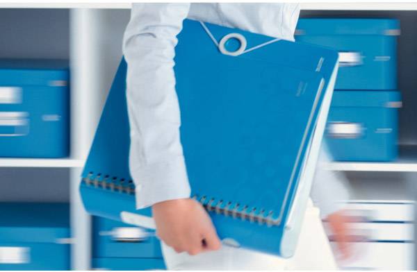 A person is carrying a large, blue ring binder; background with a shelf full of blue boxes.