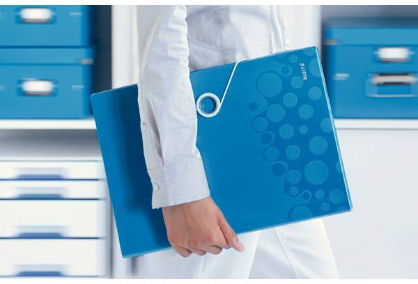 A person is holding a blue folder, wearing white clothing, and standing in front of a shelf with blue boxes.