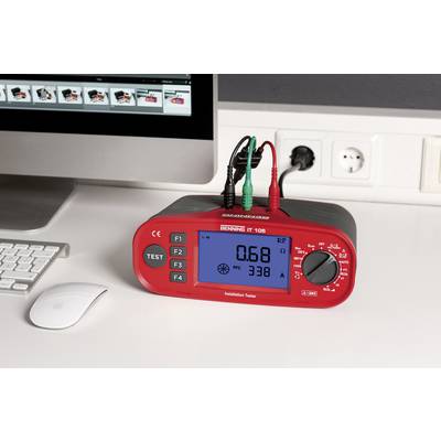 A red installation testing device with a digital display shows '0.68' and '338 A'. It is lying on a desk next to a monitor and a mouse.