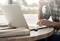 A person is typing on a laptop at a round wooden table and holding a cup. Blurred natural scenery can be seen in the background.