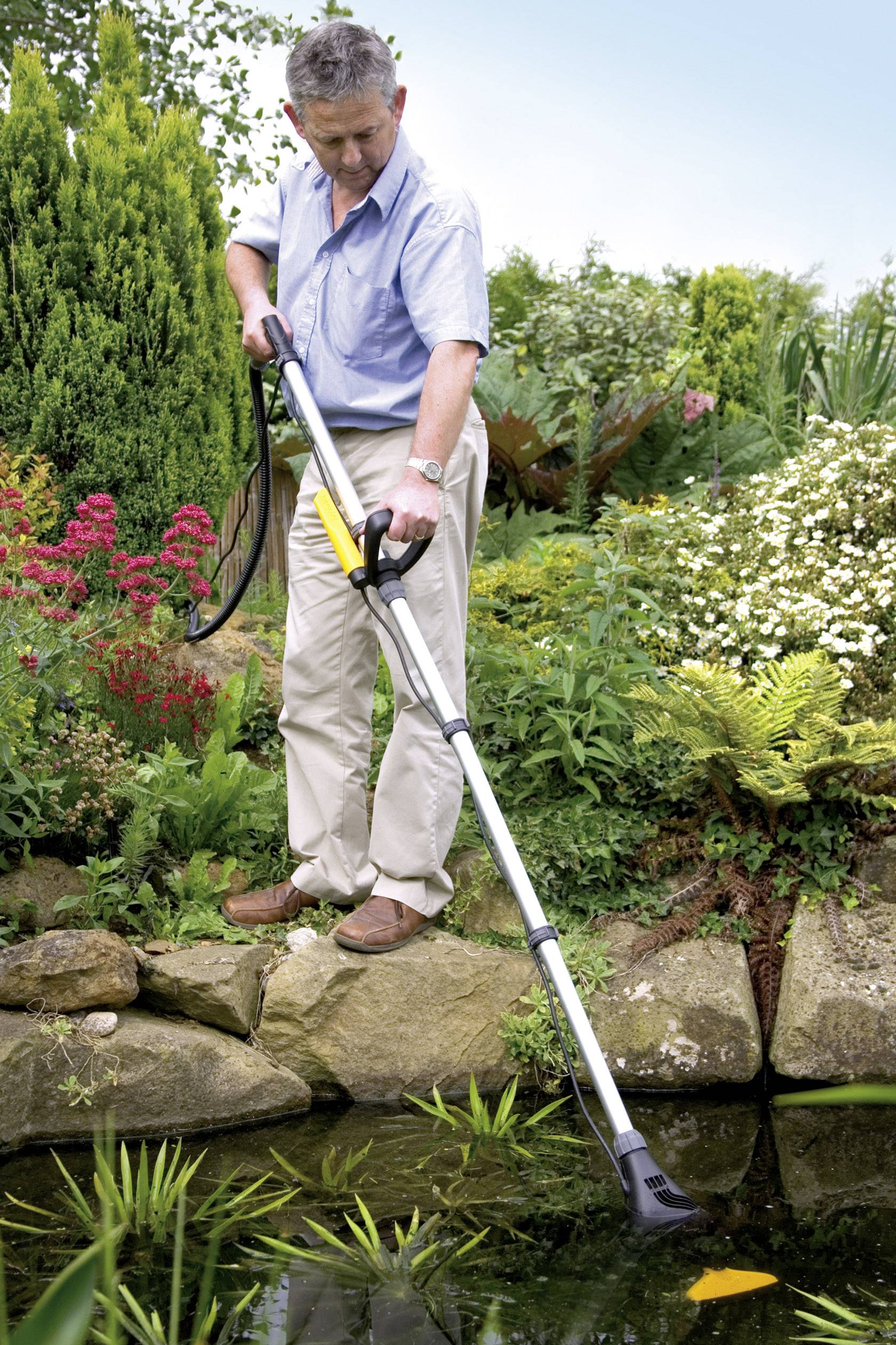 A man is cleaning a small garden pond with a pond net, surrounded by lush plants and flowers.