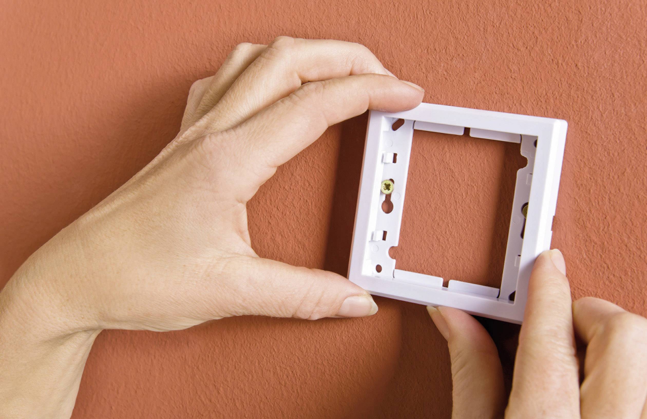 Two hands are installing a rectangular white switch frame on a red wall.