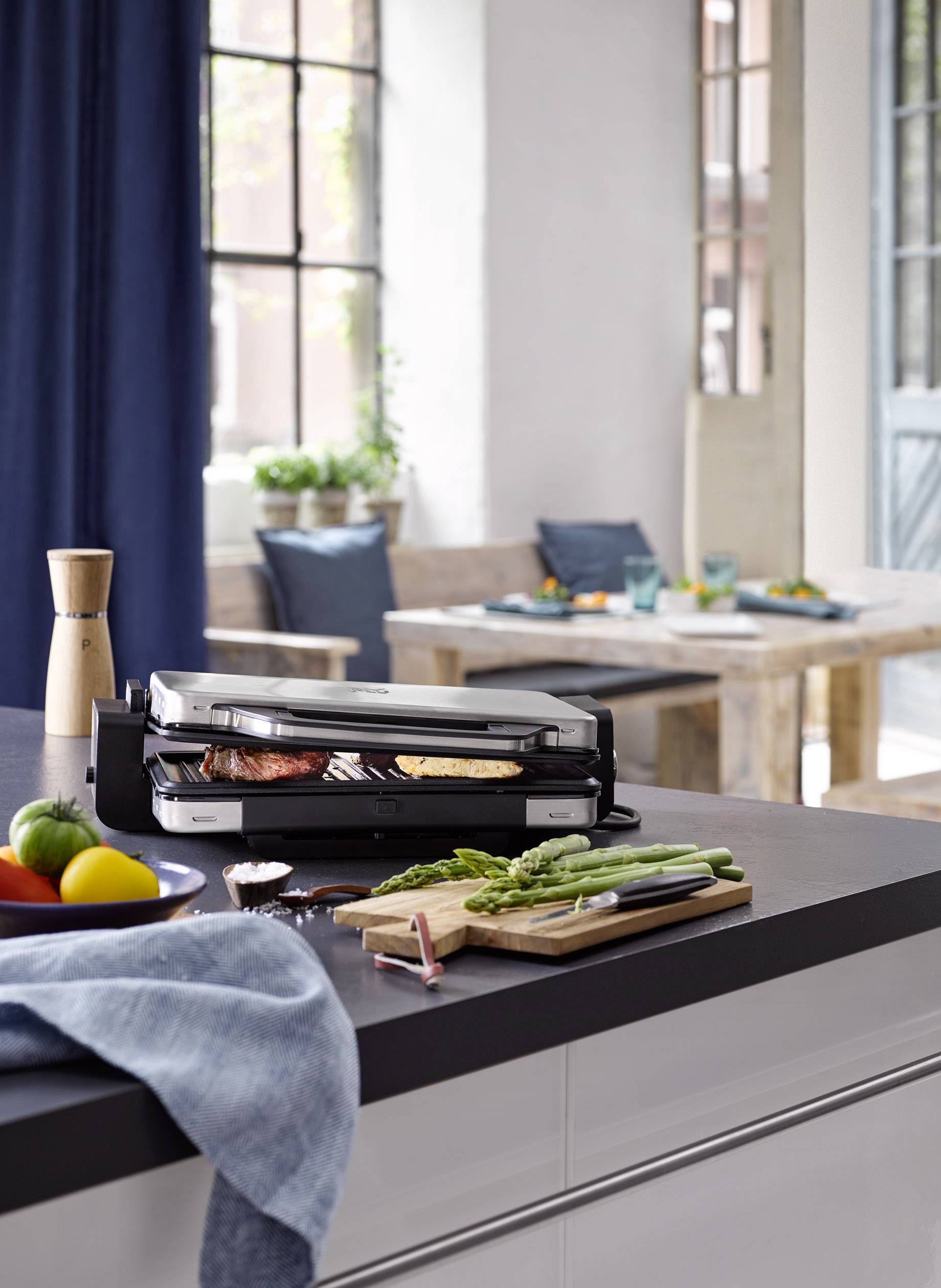 Electric grill on kitchen worktop grilling meat and vegetables. In the background, dining area with wooden table and chairs, window with blue curtains.