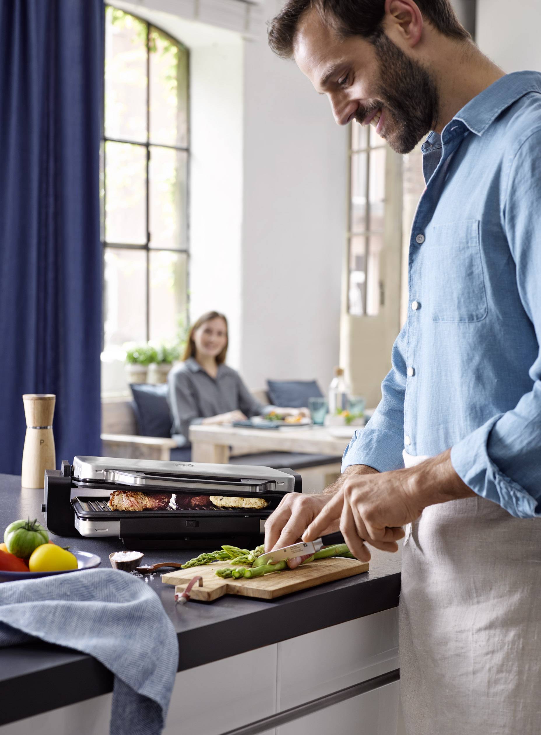A man is chopping vegetables on a chopping board in a kitchen. A woman is sitting at the table in the background.